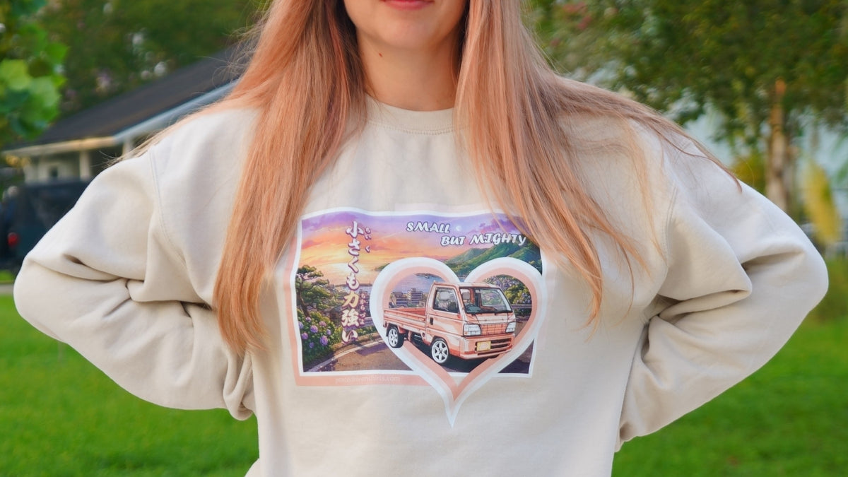 A woman wearing a cream-colored sweatshirt with a colorful graphic print of a Manga style Honda Acty inside a heart, standing outdoors on a grassy lawn.