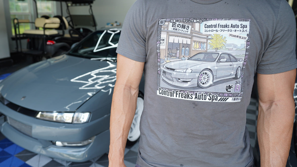 A man wearing a gray t-shirt with a graphic print of a grey s13 240sx sports car and Japanese text, standing in front of a grey s13 240sx  in a garage.